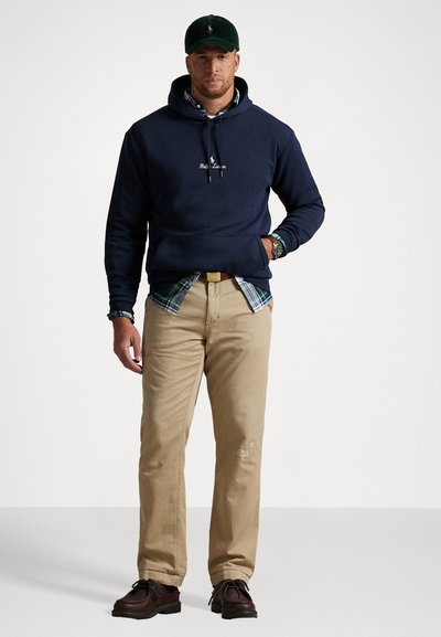 Uomo che indossa una felpa blu navy, una camicia a quadri verdi, pantaloni beige, scarpe marroni scure, un cappellino verde e un orologio da polso, in piedi davanti a uno sfondo bianco.