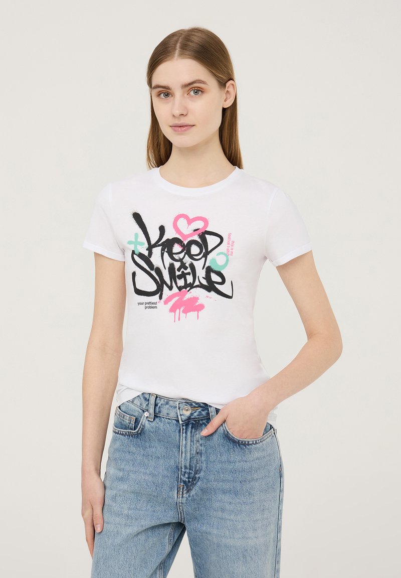 T-shirt bianca in cotone con stampa grafica contenente testo nero, rosa e verde. Design aderente con maniche corte, abbinata a jeans azzurri chiari.