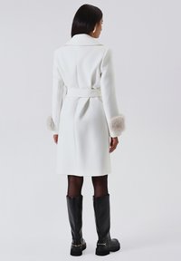Cappotto bianco in lana con vita cintura, caratterizzato da polsini in pelliccia sintetica e colletto classico. Indossato con stivali neri in pelle al ginocchio.