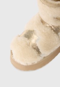 Die cremefarbene Schaffellstiefel mit einem metallisch goldenen Akzent. Das Design zeichnet sich durch eine weiche, flauschige Textur und eine runde Zehenpartie mit einer beigen Sohle aus.