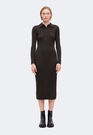 Femme debout portant une robe midi à manches longues côtelée marron foncé avec des boutons et des bottines noires sur un fond gris clair.
