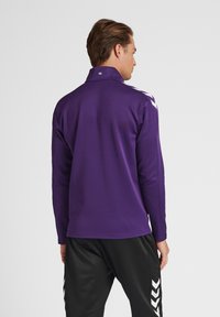 Maglietta atletica a maniche lunghe viola con colletto alto, caratterizzata da bordi bianchi lungo le spalle e una texture liscia. Visibili pantaloni atletici neri.