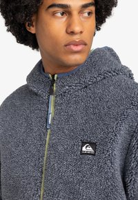 Hoodie en polaire grise avec un tissu texturé, une capuche et une fermeture éclair avant. Présente un empiècement avec logo et un détail de fermeture éclair jaune contrastant.