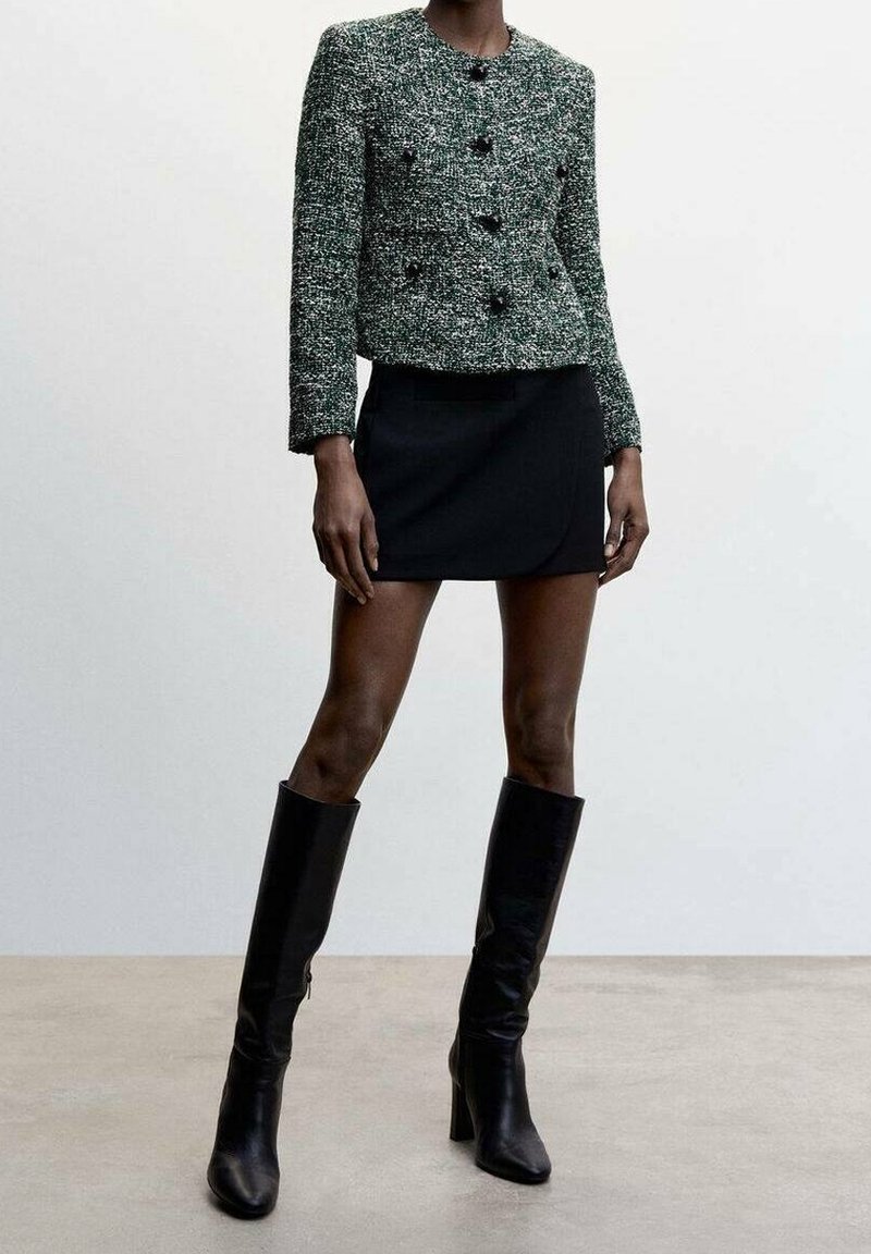 Veste en tweed verte et noire à col rond avec fermeture par boutons, associée à une mini-jupe noire et des bottes hautes en cuir noir.