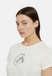 Camiseta de algodón en color blanco roto con cuello redondo y mangas cortas, que presenta bordados florales en azul y verde en la parte delantera.