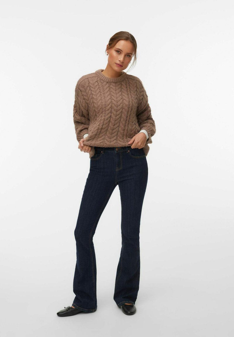 Femme debout portant un pull en maille torsadée marron, un jean évasé bleu foncé et des ballerines noires sur fond blanc.