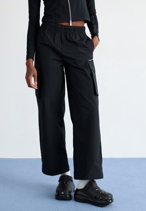 Person trägt schwarze weite Cargo-Hosen, schwarzes kurzes Zip-Up-Oberteil, weiße Socken und schwarze Plateauslipper und steht auf blauem Boden.