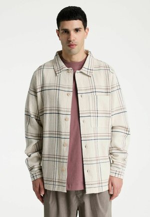 Man die een crèmekleurig, gestructureerd geruite jas draagt over een mauve overhemd en grijze broek, staand tegen een effen witte achtergrond.