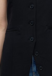 Gros plan d’un gilet noir boutonné avec deux poches fendues porté sur un jean en denim bleu.