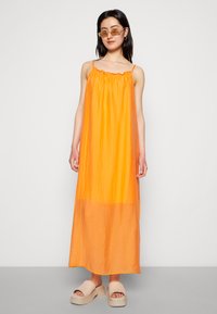 Vestido longo, sem mangas, feito de tecido leve, numa cor laranja vibrante com decote franzido. As sandálias têm solas grossas.