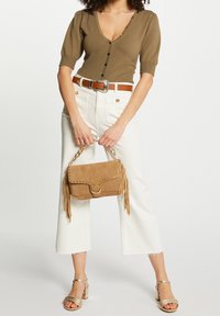 Bolso de ante marrón con detalles de flecos, una solapa curva y acentos de tachuelas. El modelo lleva una blusa ajustada de color oliva, pantalones blancos y tacones dorados con tiras.