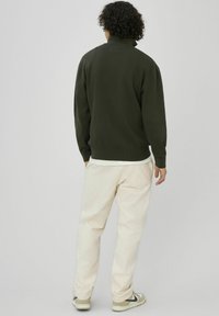 Pull tricoté vert foncé avec un col montant, porté sur une chemise blanche, associé à un pantalon en velours côtelé couleur crème et des baskets beiges.