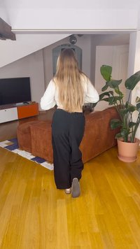 Zwarte broek met een elastische tailleband, wit cropped topje, lang steil haar, houten vloer, een bruine corduroy bank en een kamerplant.