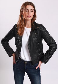 Giacca da biker in pelle nera con un design attillato, tasche con zip e dettagli imbottiti sulle maniche, abbinata a una camicia bianca e jeans.