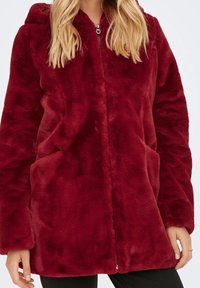 Vrouw in een bordeauxrode plush jacket met ritssluiting, lange mouwen en zijzakken, gecombineerd met zwarte broek.