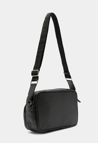 Melnā plecu soma, izgatavota no teksturēta materiāla, taisnstūra forma, regulējama siksna, ar smalkiem reljefiem detalizējumiem un sudraba aparatūru.