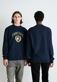 Sudadera de color azul marino con detalles del logo en la parte delantera, cuello redondo, mangas largas, y puños y dobladillo de canalé. Llevada por dos modelos.