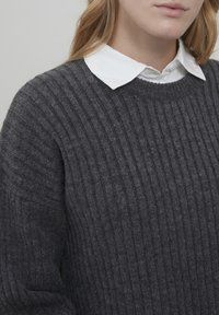 Dark gray ribbed sweater with textured knit and round neckline, layered over a white collared shirt. Close-up view of fabric details.