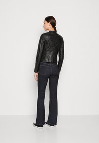 Giacca di pelle nera con un design aderente e cuciture sul retro, abbinata a jeans svasati blu scuro e sneakers bianche e nere.