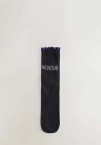 Calzino nero con polsino arricciato viola e la parola "Mercoledì" ricamata in bianco sulla parte superiore a costine, adagiato su uno sfondo bianco.