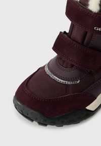Burgundi színű téli csizma, amely szintetikus és velúr anyagokból készült, texturált szövet, tépőzáras pántok, visszaverő elemek és strapabíró fekete talp jellemzi.