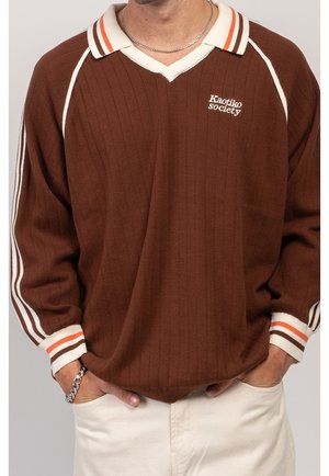 Brązowa, prążkowana koszula z długim rękawem z białym i pomarańczowym paskowanym kołnierzem i mankietami, białe logo „Kaotiko Society”, noszona z białymi spodniami.
