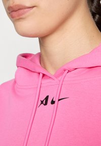 Ružová mikina s kapucňou a šnúrkou, s čiernym logom Nike na hrudi. Tkanina sa zdá byť jemná s hladkou textúrou.