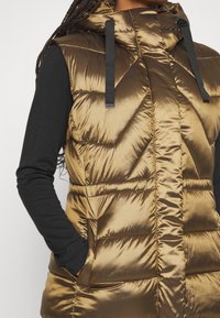 Ärm-lös, huva-puffer väst i metalliskt brons med chevron quiltad textur. Har svarta snoddar och sidofickor.