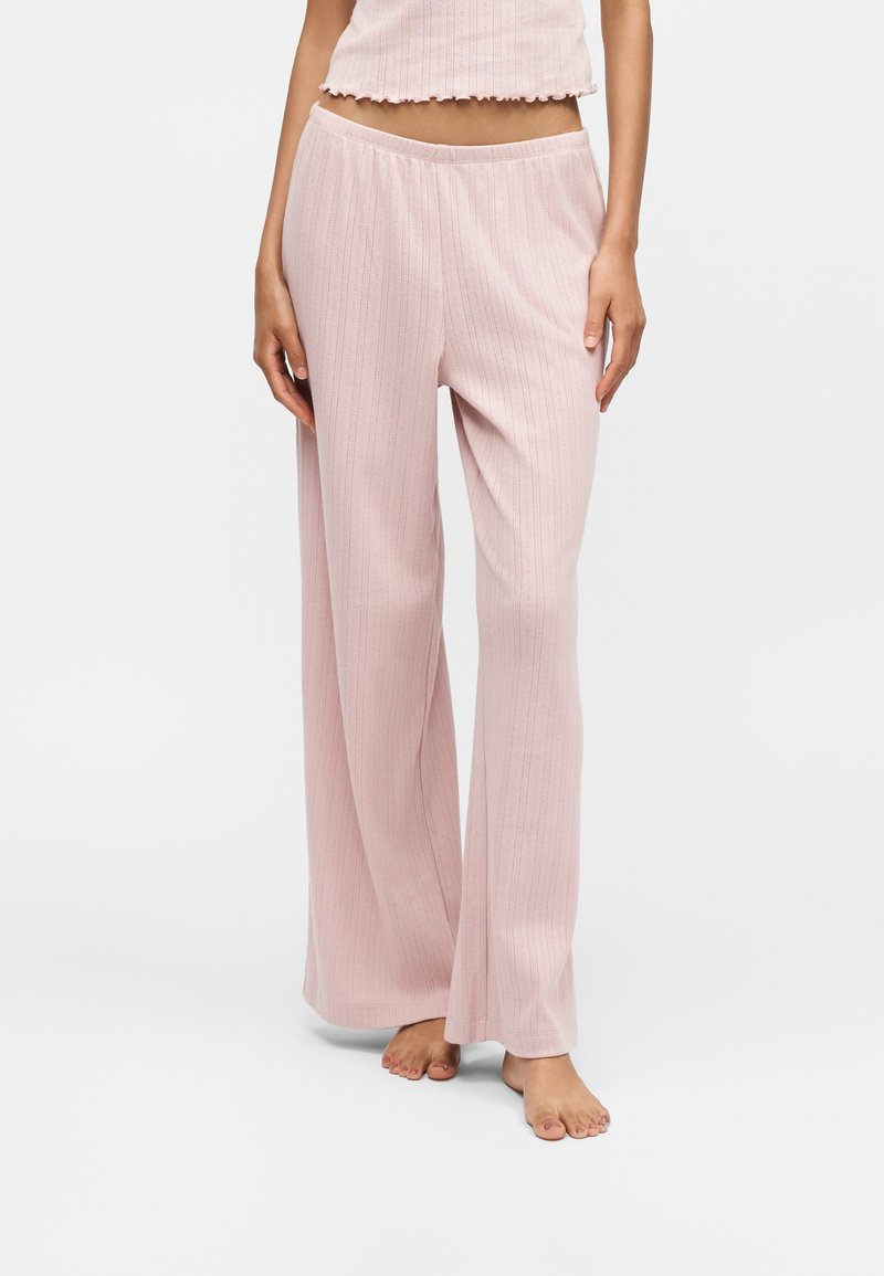 Femme portant un pantalon large côtelé rose clair et un top court assorti, debout pieds nus sur fond blanc.