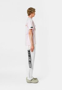 Camisa de manga corta rosa con estampado gráfico, combinada con joggers blancos que presentan una etiqueta negra vertical. Unas zapatillas grises de suela gruesa completan el look.