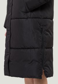 Abrigo tipo puffer negro con un diseño acolchado, que cuenta con un zipper lateral. La tela es mate y la forma general es larga con un corte recto.