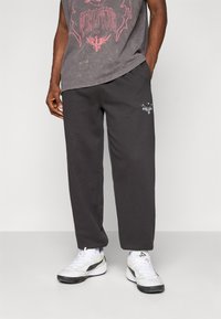 Mand i sorte sweatpants med et lille hvidt phoenix-logo på venstre lår og hvide sportsko, stående med hænderne i lommerne.