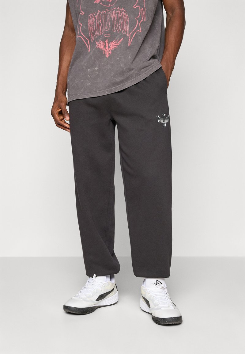Mand i sorte sweatpants med et lille hvidt phoenix-logo på venstre lår og hvide sportsko, stående med hænderne i lommerne.