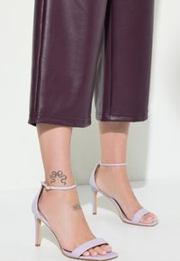 Burgundské cropped kalhoty z umělé kůže, které jsou kombinovány se světle fialovými sandály na vysokém podpatku s řemínky kolem kotníků a otevřenou špičkou. Viditelné dekorativní tetování.