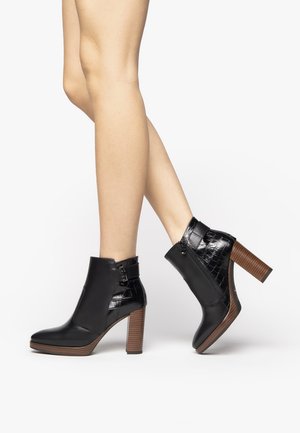 Botas negras hasta el tobillo con una textura brillante de cocodrilo en la parte trasera, tacón alto y apilado, y detalle de hebilla en el lateral. Acabado en cuero suave.