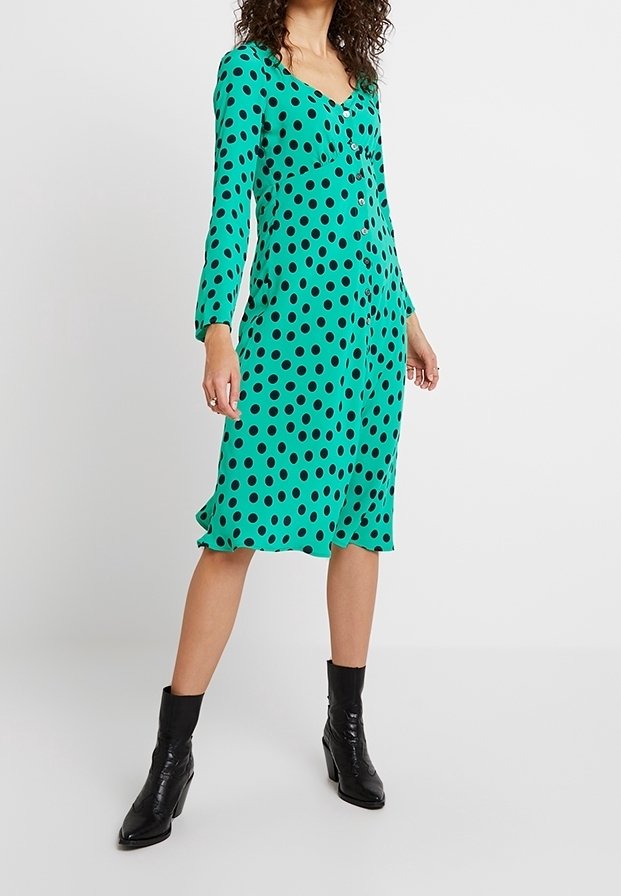Femme portant une robe verte à manches longues, boutonnée, avec des pois noirs, et des bottines noires, debout devant un fond blanc.