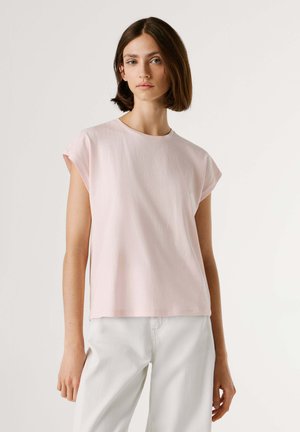 Mujer con cabello corto y castaño lleva una camiseta de manga corta rosa claro y pantalones blancos, de pie frente a un fondo claro y liso.