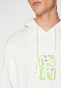 Sweat-shirt à capuche blanc en coton, orné d'un motif vert stylisé avec le mot « love » et des accents de fleurs roses à l'avant.