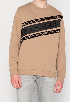 Sudadera beige con banda negra brillante que presenta el texto "GLORIOUS" y un patrón griego. Puños y dobladillo acanalados, material suave.