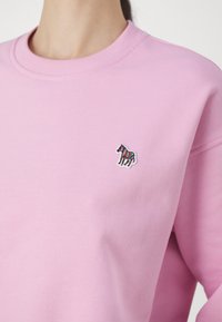 Růžová mikina vyrobená z měkké tkaniny, s malým vyšitým barevným logem zebry na hrudi a klasickým kulatým výstřihem.