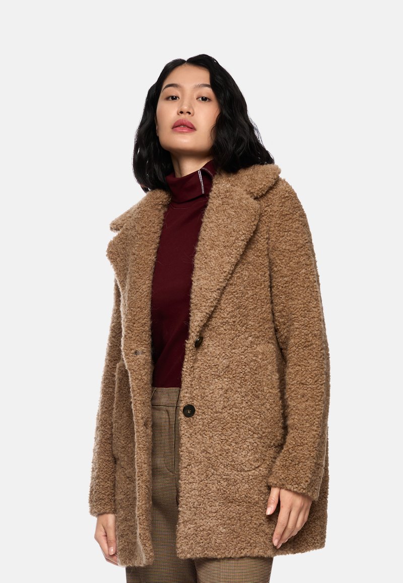 Brauner strukturierter Mantel mit Kragen, zwei Knöpfen und Seitentaschen. Trägt über einem burgunderfarbenen Rollkragenpullover und karierter Hose.