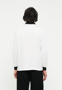 Camisa de manga larga en algodón blanco con cuello y puños negros. Presenta un diseño simple con un dobladillo recto y un ajuste relajado.