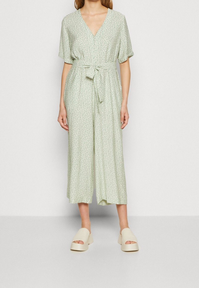 Femme portant une combinaison vert clair à manches courtes avec un petit imprimé floral, col en V, taille nouée, jambes larges raccourcies et sandales à plateforme beiges.