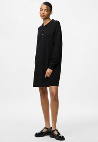 Melnā adīta kleita ar garām piedurknēm, polo apkakli, trīs pogu atvērumu un brīvu piegriezumu, kas savienota ar spīdīgām melnām siksnu kurpēm.