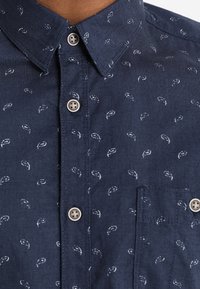 Granatowa koszula zapinana na guziki z małymi białymi wzorami paisley i kieszenią na piersi, noszona przez osobę.