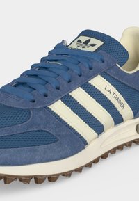 Μπλε αθλητικό παπούτσι Adidas L.A. Trainer με λευκές ρίγες, επάνω μέρος από καστόρι και δίχτυ, υφή στη σόλα και λογότυπο Adidas στη γλώσσα.
