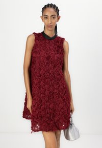 Bordeaux jurk van gestructureerde stof met roos appliqués, met een mouwloze ontwerp en een zwarte kraag. Een kleine zilveren handtas wordt vastgehouden.