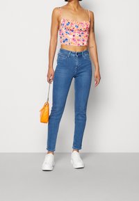 Różowa kwiatowa bluzka na ramiączkach, zestawiona z niebieskimi dopasowanymi dżinsami. Trzyma małą pomarańczową torbę, nosi białe sneakersy.