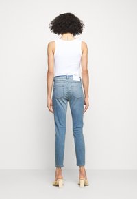 Jeans de mezclilla azul claro con un ajuste slim, cintura media y dos bolsillos traseros; combinados con una blusa sin mangas blanca y sandalias de tacón cuadrado beige.