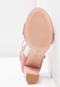 Sandale à talons hauts en suede rose avec un dos ouvert et un design à brides, présentant une semelle texturée de couleur marron clair et un détail de semelle intérieure avec logo.